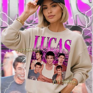 May include: Beige sweatshirt featuring a graphic of Lucas Scott in a vintage style. The design includes the name "LUCAS" in pink, with multiple images of the actor. The sweatshirt is worn with a black cap and gold bracelets.