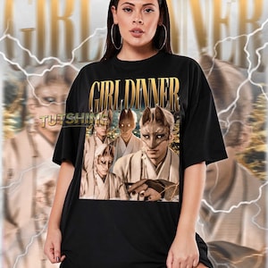 May include: Black unisex t-shirt with the text "GIRL DINNER" in gold lettering. The shirt features a graphic of people wearing fox masks. The brand name "TUTSHINE" is also visible. The model is wearing a 2XL shirt.