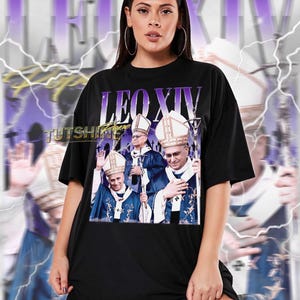 Könnte beinhalten: Schwarzes Unisex-T-Shirt mit einer Grafik von drei Figuren in religiöser Kleidung und dem Text "LEO XIV" in Lila. Das Shirt hat einen Vintage-Stil mit verschwommenem Hintergrund. Der Text "TUTSHINE" ist ebenfalls sichtbar. Das Shirt ist größengetreu.