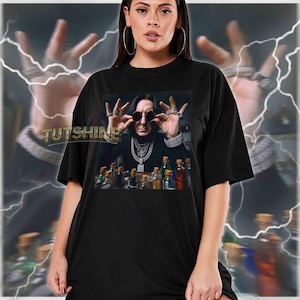 May include: Black unisex t-shirt featuring a graphic of a person wearing sunglasses and jewelry, with a collection of bottles in front. The shirt has the text "TUTSHINE" on the front. The model is wearing a size 2XL.