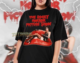 Puede incluir: Camiseta unisex negra con el texto rojo "THE ROCKY HORROR PICTURE SHOW" y una imagen de una mujer sobre labios rojos. La camiseta tiene mangas cortas y cuello redondo. La modelo lleva una talla 2XL.