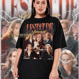 May include: Black unisex t-shirt featuring a graphic of the word "LESTAT DE Lioncourt" in orange and yellow, with a collage of images of a man. The shirt is worn by a person. The text "TUTSHINE" is also visible.