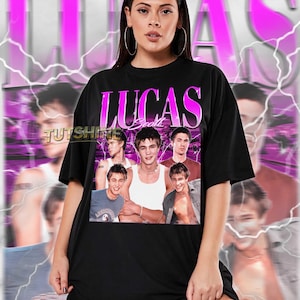 May include: Black unisex t-shirt with a pink and white "LUCAS Scott" design and a collage of images. The shirt has a crew neck and short sleeves. The text "TUTSHINE" is also visible. The shirt is worn by a person.