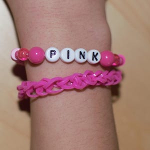 May include: A pink beaded bracelet with the word "PINK" spelled out in white beads. The bracelet is worn on a person's wrist.