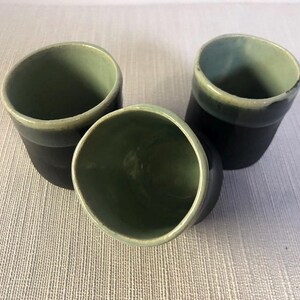 Peut inclure: Trois tasses en céramique avec un extérieur noir et un intérieur vert. Les tasses sont disposées en forme de triangle.