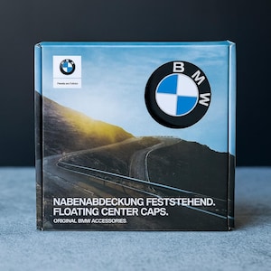 May include: Box of BMW floating center caps. The box features the BMW logo and a scenic road image. Text on the box reads "NABENABDECKUNG FESTSTEHEND. FLOATING CENTER CAPS. ORIGINAL BMW ACCESSORIES."