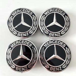 Op de afbeelding: Vier Mercedes Benz wieldopjes. Elke dop heeft een zwart midden met een zilveren Mercedes-Benz logo en de woorden "Mercedes" en "Benz" in zilveren letters. De doppen hebben een zilveren buitenring met een laurierkrans ontwerp.