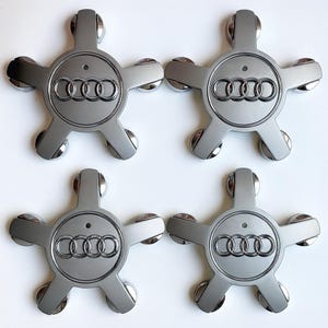 May include: Four silver-gray wheel center caps with the Audi logo. Each cap has a star-like design with five arms and a circular center. The Audi logo is in the center of each cap.