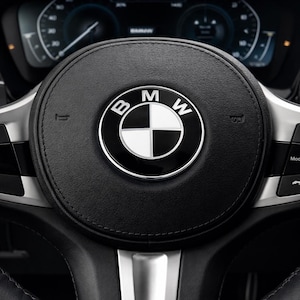 Könnte beinhalten: Nahaufnahme eines schwarzen Lederlenkrads mit dem BMW-Logo in der Mitte. Das Logo ist ein schwarzer Kreis mit silbernem Rand, und das BMW-Emblem befindet sich in der Mitte. Der Hintergrund zeigt das Armaturenbrett des Autos.