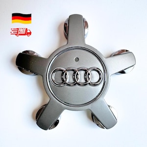 May include: A silver-colored Audi wheel center cap with a five-spoke design. The Audi logo is prominently displayed in the center. The cap has a metallic finish and a glossy, reflective surface. The image also includes a German flag and a free delivery icon.