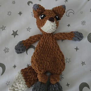 May include: A handmade, plush fox toy. The fox is primarily brown with a white chest and tail tip, and gray accents on the ears, paws, and feet. The toy has black eyes and a brown nose. The fox is made of a soft, textured yarn.