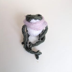 May include: A knitted frog ornament with a white body, grey head and limbs, and a lilac scarf. The frog's limbs are extended, and the design is whimsical. The frog is a charming handmade item.