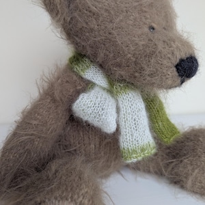 May include: A handmade teddy bear with a soft, brown, fuzzy texture. The bear has a black nose and is wearing a white and green scarf. The bear is a plush toy.