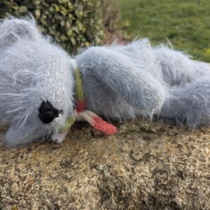 May include: A handmade, light blue plush koala toy with a black eye and a colorful scarf. The koala is lying on a rock, with its fluffy fur and small ears visible. The scarf has green, red, and white stripes.