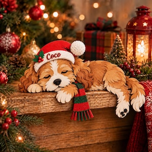 Può includere: Una sagoma in legno a strati raffigurante un cane spaniel marrone e bianco che dorme, con un cappello da Babbo Natale rosso con il nome Coco scritto in corsivo e una sciarpa a righe verdi e rosse, appoggiato su una superficie in legno con sfondo festivo.