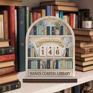 May include: A wooden bookshelf-shaped decorative piece with the text "BEACH READS & SUNSHINE" and "NANA'S COASTAL LIBRARY". It features miniature books in various colors, floral accents, and number tiles. The numbers displayed are 1, 4, and 6.