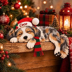 Könnte beinhalten: Ein mehrschichtiger Holzausschnitt eines schlafenden Hundes mit Merle-Fell, der eine rote Weihnachtsmütze mit dem in weißer Schreibschrift gestickten Namen Coco sowie einen grün-rot gestreiften Schal trägt. Der Hund liegt auf einer Holzoberfläche.