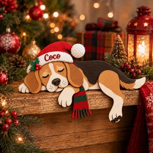 May include: A wooden cutout of a sleeping beagle dog wearing a red Santa hat with the name Coco written in script and a green and red striped scarf. The dog has brown, white, and black fur markings and is resting with its eyes closed.