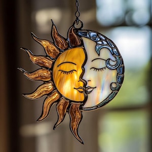 Könnte beinhalten: Hängende Dekoration aus Buntglas mit Sonne und Mond. Die Sonne ist warmgelb und goldfarben, mit detaillierten Strahlen. Der Mond ist perlmuttweiß und blau. Die beiden Figuren sind mit geschlossenen Augen an den Lippen verbunden.
