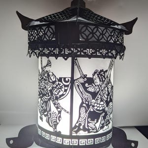 May include: A black metal lantern with a white paper cylinder inside. The cylinder features a black silhouette of two figures fighting with swords. The lantern has a decorative top with a black and white pattern.