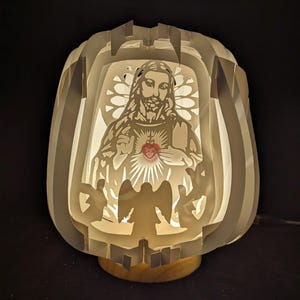 Może przedstawiać: Biała papierowa lampa z warstwowym wzorem przedstawiającym wycięty obraz Jezusa z sercem. Lampa jest podświetlana od wewnątrz, dając ciepłe światło. Podstawa lampy jest drewniana.