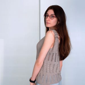 May include: A sleeveless, light brown knitted top with a decorative openwork pattern. The top has a scoop neck and a slightly fitted silhouette. The model is wearing glasses and a black watch.