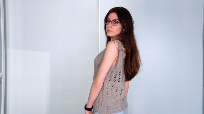 May include: A sleeveless, light brown knitted top with a decorative openwork pattern. The top has a scoop neck and a slightly fitted silhouette. The model is wearing glasses and a black watch.