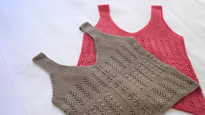 May include: Two knitted tank tops in the image. One is a muted rose color, and the other is a taupe color. The taupe tank top has a detailed openwork design. The tank tops are lying on a white surface.