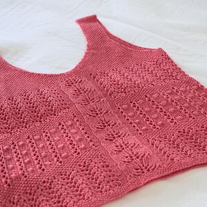 May include: A dusty rose-colored knitted tank top with a scoop neck and shoulder straps. The top features intricate openwork patterns, including vertical lines, leaf-like designs, and small bobbles, creating a textured appearance. The garment is displayed on a white surface.