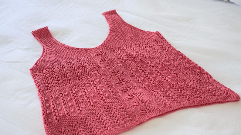 May include: A dusty rose-colored knitted tank top with a scoop neck and shoulder straps. The top features intricate openwork patterns, including vertical lines, leaf-like designs, and small bobbles, creating a textured appearance. The garment is displayed on a white surface.