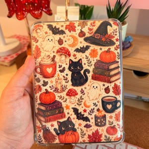 Könnte beinhalten: Eine rechteckige Tablethülle mit einem herbstlichen Muster aus schwarzen Katzen, Geistern, Kürbissen, Pilzen, Bücherstapeln, Fledermäusen, Kerzen und Tassen in Orange-, Rot-, Braun- und Schwarztönen auf einem cremefarbenen Hintergrund.