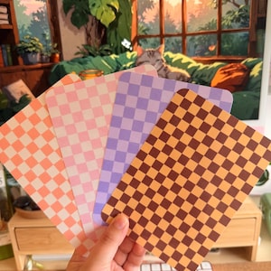 Könnte beinhalten: Ein Set aus vier rechteckigen Karten mit einem sich wiederholenden Schachbrettmuster in verschiedenen Farben. Die Karten sind fächerförmig angeordnet und zeigen Farbtöne in Orange, Hellrosa, Lavendel sowie ein dunkelbraun-beiges Karomuster.