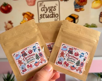 May include: Three brown paper pouches, each featuring a rectangular sticker with a festive holiday theme. The stickers display illustrations of a teddy bear, snowman, gingerbread man, and Christmas tree, with the text Christmas Suprise Stickers.