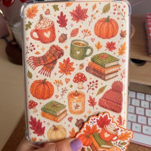 Könnte beinhalten: Eine transparente Schutzhülle für einen E-Reader mit einem herbstlichen Muster aus orangefarbenen Kürbissen, grünen und roten Tassen, Bücherstapeln, bunten Herbstblättern, Eicheln, Pilzen, einem Strickschal, einer Mütze und einer brennenden Kerze.