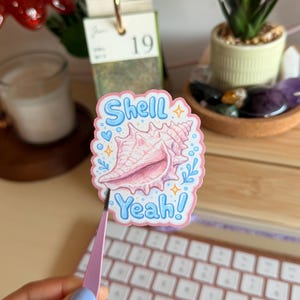 Könnte beinhalten: Ein gestanzter Aufkleber mit einer Illustration einer rosa Muschel, zentriert auf weißem Hintergrund mit hellrosa Rand. Der Text Shell Yeah! steht in blauen Blasenbuchstaben über und unter der Muschel, akzentuiert durch kleine blaue Herzen und Sterne.