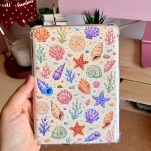 Könnte beinhalten: Eine transparente Schutzhülle für Tablets mit einem sich wiederholenden Aquarellmuster aus verschiedenen Muscheln, Seesternen und Algen in Rosa-, Lila-, Orange-, Blau- und Grüntönen auf einem hellcremefarbenen Hintergrund.