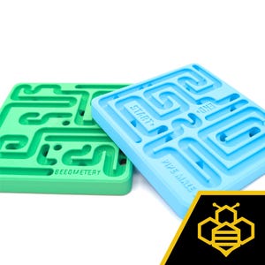 May include: Two square mazes, one green and one blue. The green maze has the word "BEEOMETRY" and the blue maze has "PIPE MAZE", "START" and "END" labels. The mazes are made of a solid material and are set against a white background.