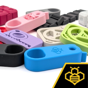 May include: A collection of colorful silicone bottle stoppers in various shades, including pink, purple, green, and blue. Each stopper features a unique design and a loop for easy removal. The blue stopper has a graphic of a bee.