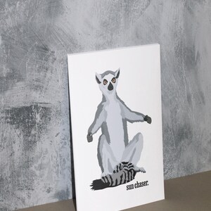 May include: A white rectangular print featuring a gray lemur illustration with orange eyes and the text "sun chaser." The lemur is sitting with its arms outstretched. The background is a textured gray wall.