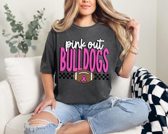 Puede incluir: Camiseta gris oscuro con el texto "pink out BULLDOGS" en blanco y rosa. Un balón de fútbol con una cinta rosa está centrado debajo del texto. La camiseta se usa con vaqueros azules.