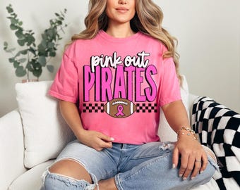 Puede incluir: Camiseta rosa con el texto "Pink Out Pirates" en blanco y rosa. Un balón de fútbol con una cinta rosa está centrado debajo del texto. La camiseta es de manga corta y cuello redondo.