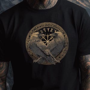 Könnte beinhalten: Schwarzes T-Shirt mit einem detaillierten kreisförmigen Design, das zwei sich gegenüberstehende Raben zeigt. Das Design enthält Runen und ein zentrales Sternmuster. Das Shirt ist aus dunklem Stoff.