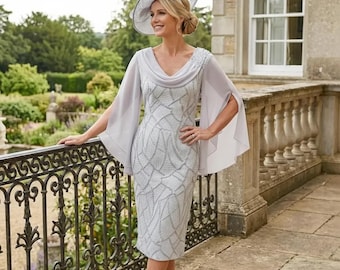 May include: A light gray knee-length dress with a cowl neckline and embellished details. The dress features a unique pattern and is paired with a matching hat and sheer cape sleeves. The outfit is suitable for formal events.
