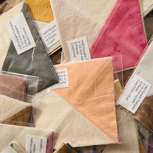 May include: Assortment of fabric squares in various colors, including peach, pink, brown, and gray. Each square is labeled with the type of fabric, color, and size. The fabric squares are packaged in clear plastic bags. The text on the labels reads: "Half-Square Triangles", "5 1/2-inch square", "6 Pieces", "Mixed Colors", "Cochineal + Logwood", "Achiot", "Cochineal", "Cutch", "Curly Dock + Rust", "Hollyhocks".
