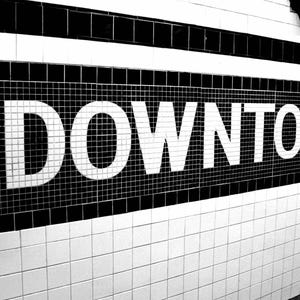Puede incluir: Un letrero de pared hecho de azulejos blancos y negros que muestra la palabra DOWNTOWN en letras mayúsculas blancas, negritas y sin remates. El texto está sobre un fondo rectangular negro de azulejos pequeños, bordeado por azulejos blancos.