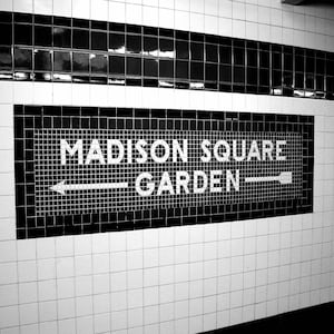 May include: A black and white subway wall sign featuring the text MADISON SQUARE GARDEN in bold capital letters. The sign is composed of small square tiles with a horizontal arrow pointing in both directions, set against a white tiled wall.
