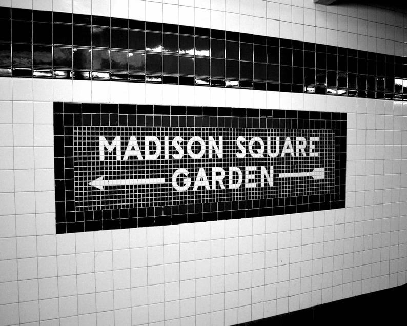 K&ouml;nnte beinhalten: Ein schwarz-wei&szlig;es U-Bahn-Wandschild mit dem Text MADISON SQUARE GARDEN in fetten Gro&szlig;buchstaben. Das Schild besteht aus kleinen quadratischen Fliesen mit einem horizontalen Pfeil, der in beide Richtungen zeigt, vor einer wei&szlig;en Fliesenwand.