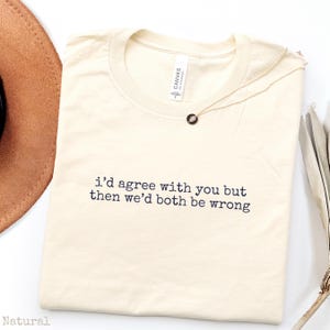 May include: A natural-colored crew neck t-shirt featuring the black printed text "i'd agree with you but then we'd both be wrong" centered on the chest. The shirt has a standard short-sleeve cut and a visible brand label inside the neckline.