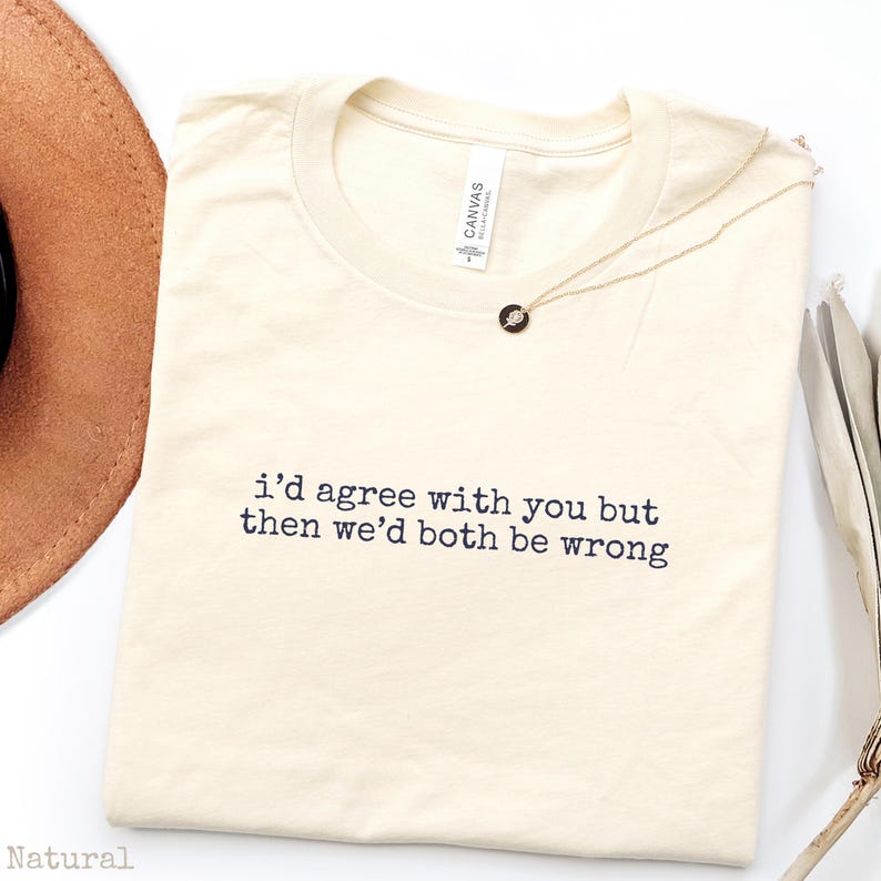 May include: A natural-colored crew neck t-shirt featuring the black printed text "i'd agree with you but then we'd both be wrong" centered on the chest. The shirt has a standard short-sleeve cut and a visible brand label inside the neckline.