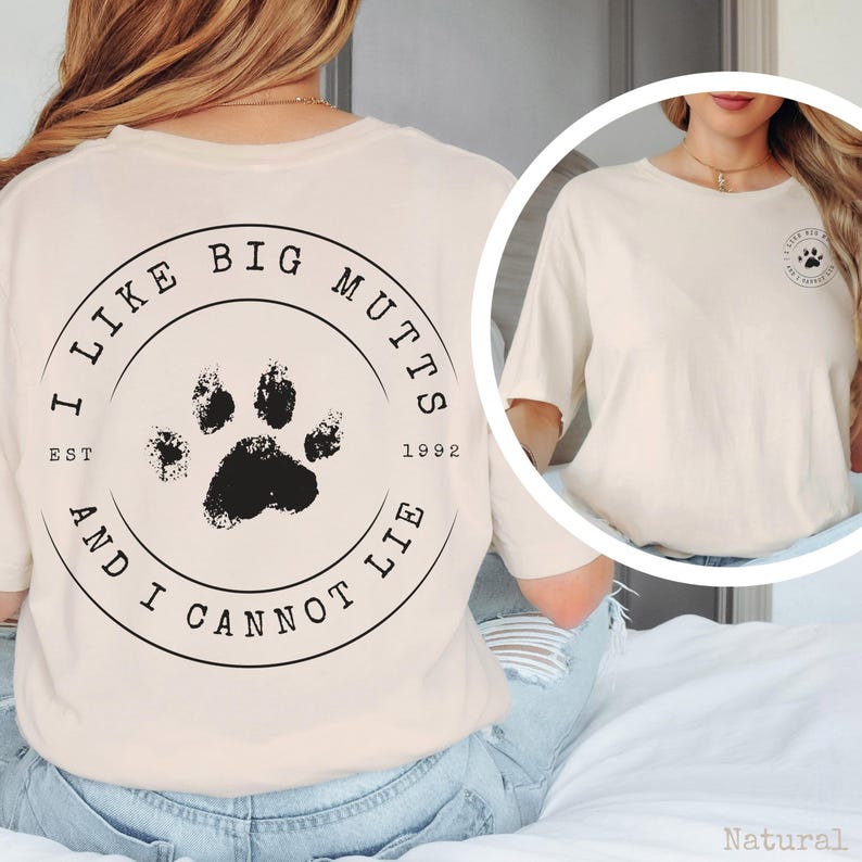 K&ouml;nnte beinhalten: Ein naturfarbenes T-Shirt mit einer schwarzen kreisf&ouml;rmigen Grafik auf der R&uuml;ckseite, die einen Pfotenabdruck in der Mitte und den Text I LIKE BIG MUTTS AND I CANNOT LIE EST 1992 zeigt. Auf der Brust befindet sich eine kleinere Version der Grafik.